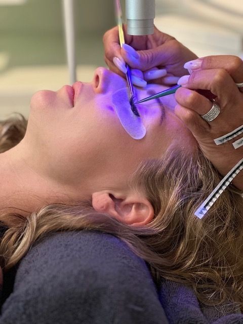 Person receiving a cosmetic eyelash treatment under blue light, with hands carefully applying extensions.